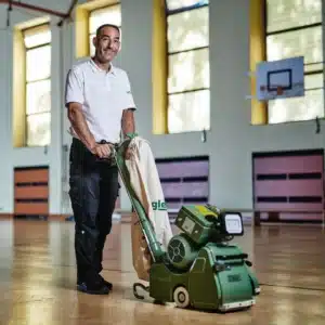 Fachmann mit Bandschleifmaschine bei substanzschonender Parkettaufbereitung in Sporthalle
